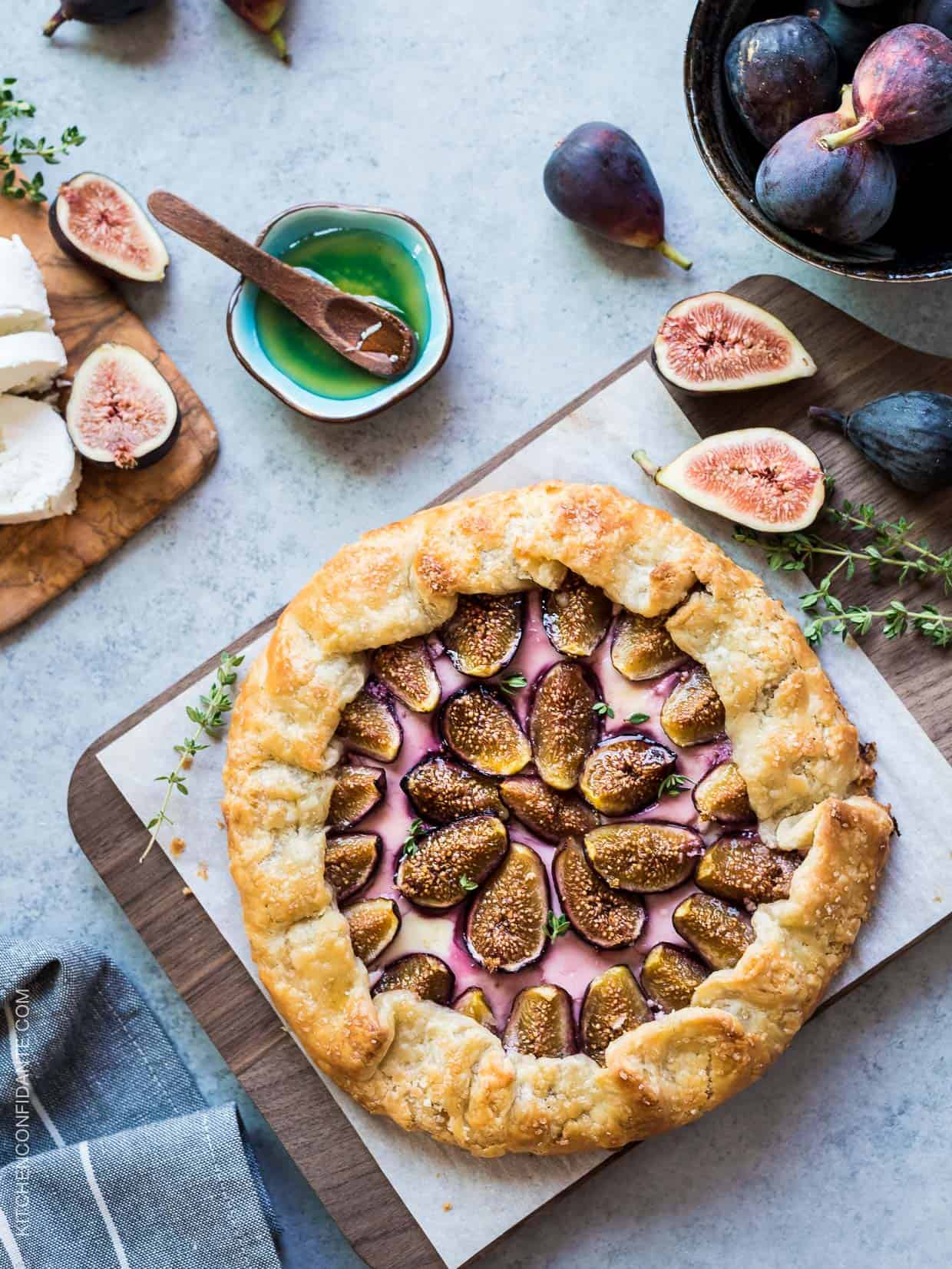 Tablescape of freshly baked Fig, Honey and Goat Cheese Galette surrounded by fresh herbs, figs, and a bowl of honey with a wooden spoon.