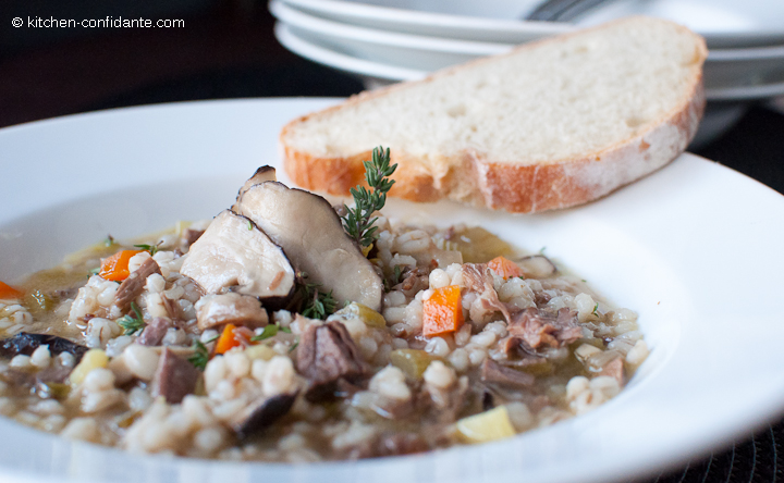 A large shallow white bowl full of beef, mushroom, and barley soup. A piece of sliced french bread sits on the edge of the bowl.