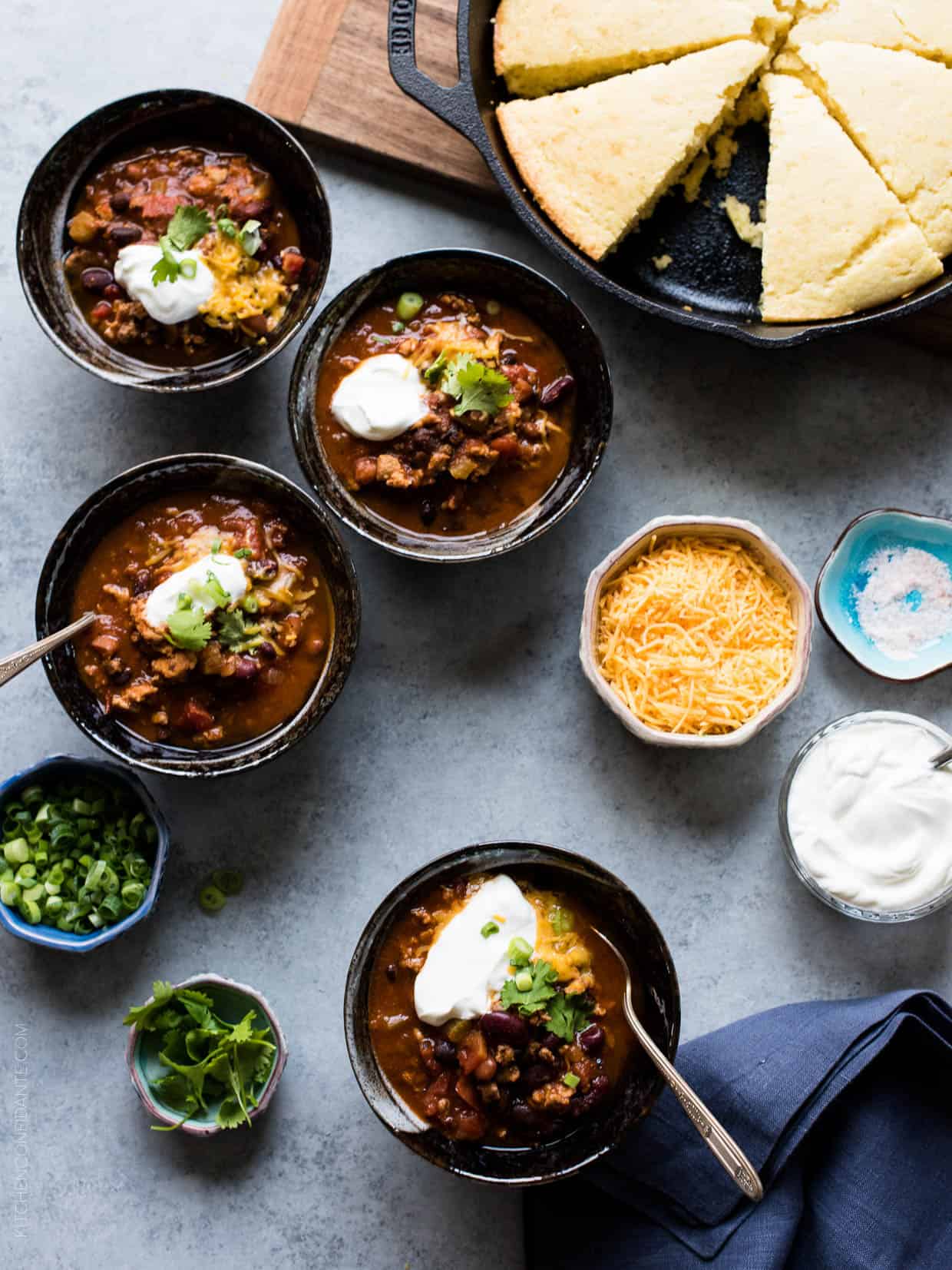 Four bowls of Classic Chili garnished with shredded cheese, sour cream, green onions and cilantro with cornbread muffins in the background. 