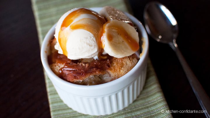 A ramekin of bread pudding topped by a small scoop of icecream, slices of banana, and drizzle of caramel sauce. The white ramekin sits on a green cloth napkin, with a spoon lying beside it.