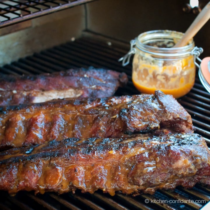 Three racks of baby back ribs on the grill with a jar of fiery peach bbq sauce.