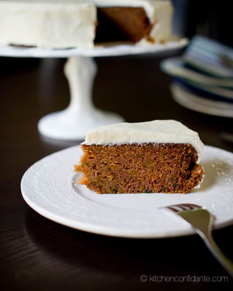 A slice of sweet potato cake on a white plate in the foreground, with the rest of the cake on a cake stand in the background.