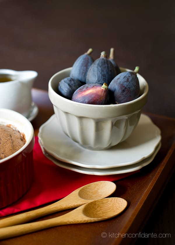 A bowl of figs sits on top of two white saucers, with two wooden spoons in the foreground.
