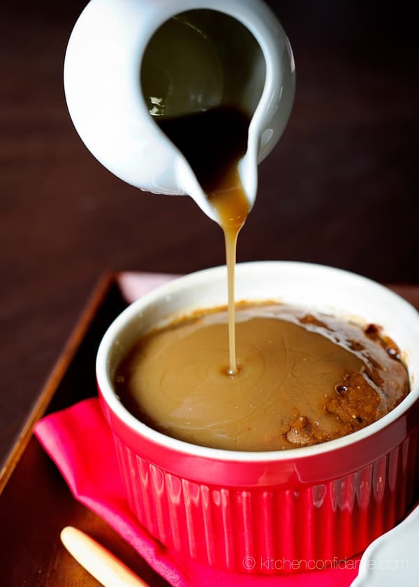 A red ramekin filled with a fresh fig cake. A small white pitcher pours toffee sauce over the top.