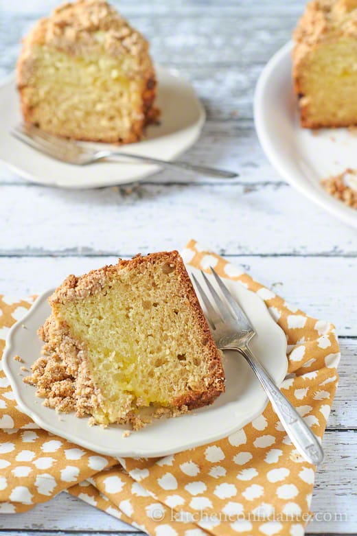 A slice of meyer lemon coffee cake on a small white plate with a fork, on an orange and white cloth.