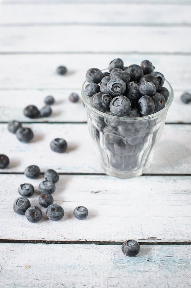 A clear glass full of fresh blueberries, with additional blueberries scattered around on the table.