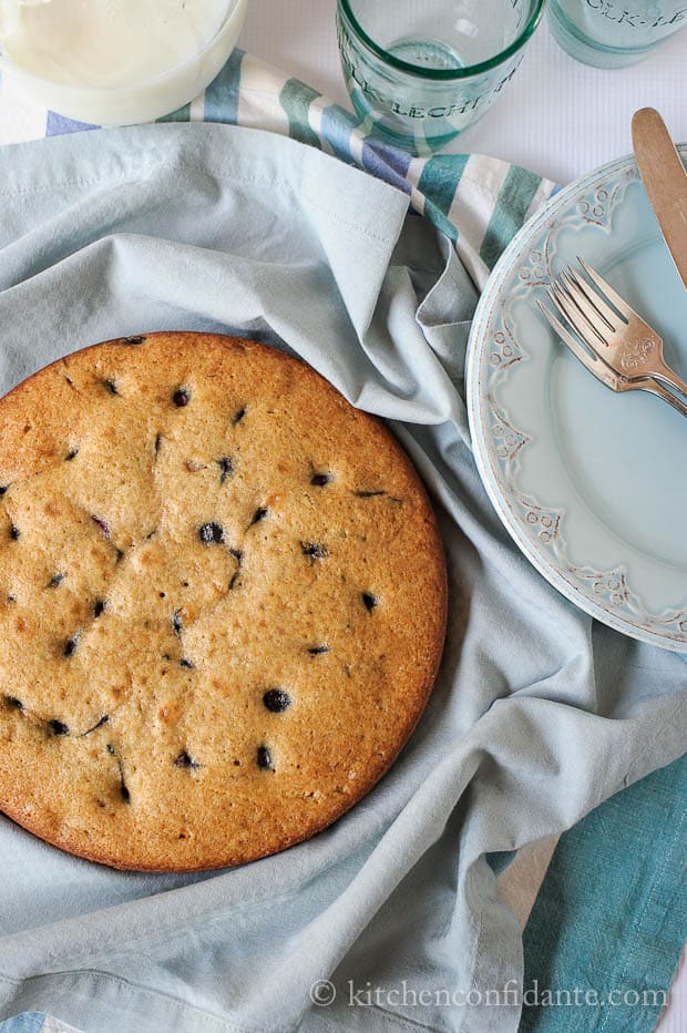 An uncut round blueberry cake sits on various blue cloths. Beside the cake is a plate, two forks, a knife, and empty glasses.