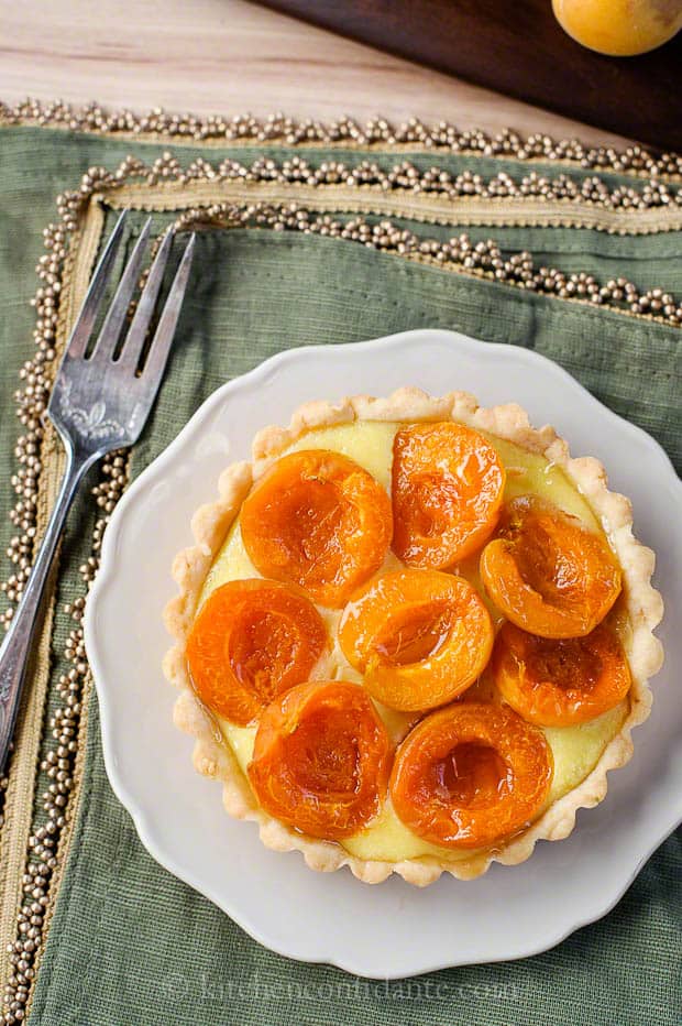 An apricot tart sits on a round decorative white plate that rests on a green napkin.