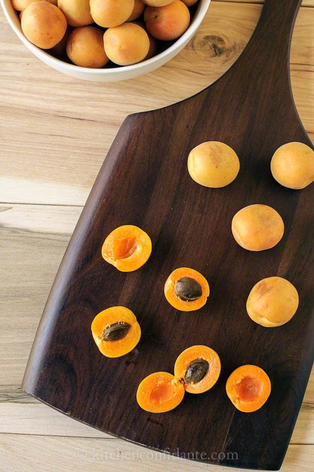Apricots sit on a dark walnut cutting board - some whole, others halved.