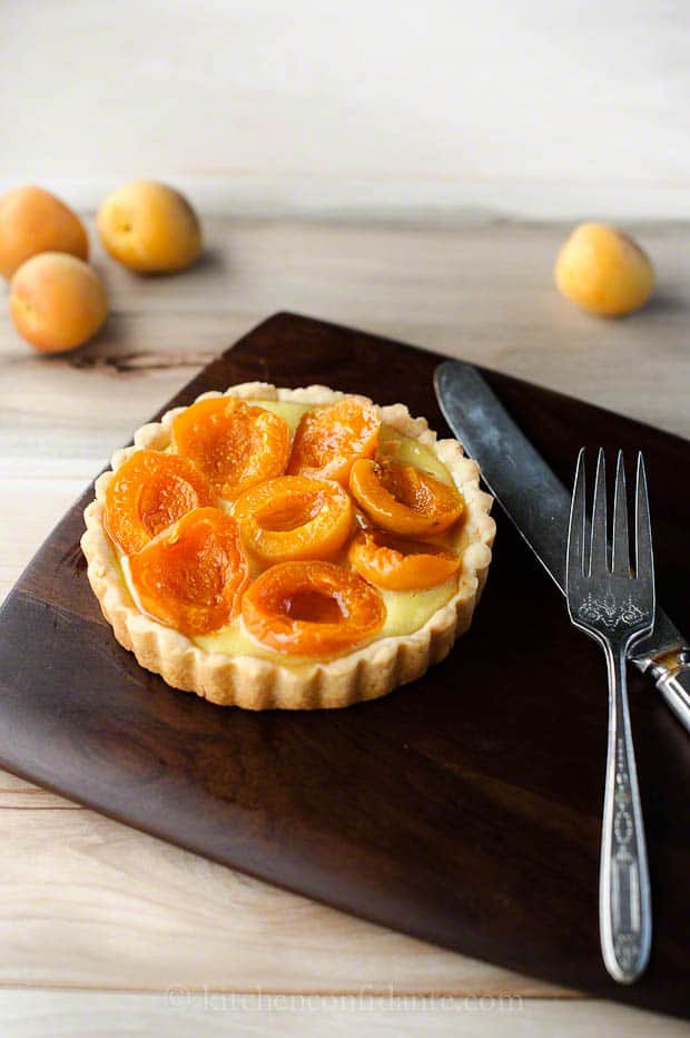 An apricot tart ready to eat sits on a cutting board with a knife and fork.