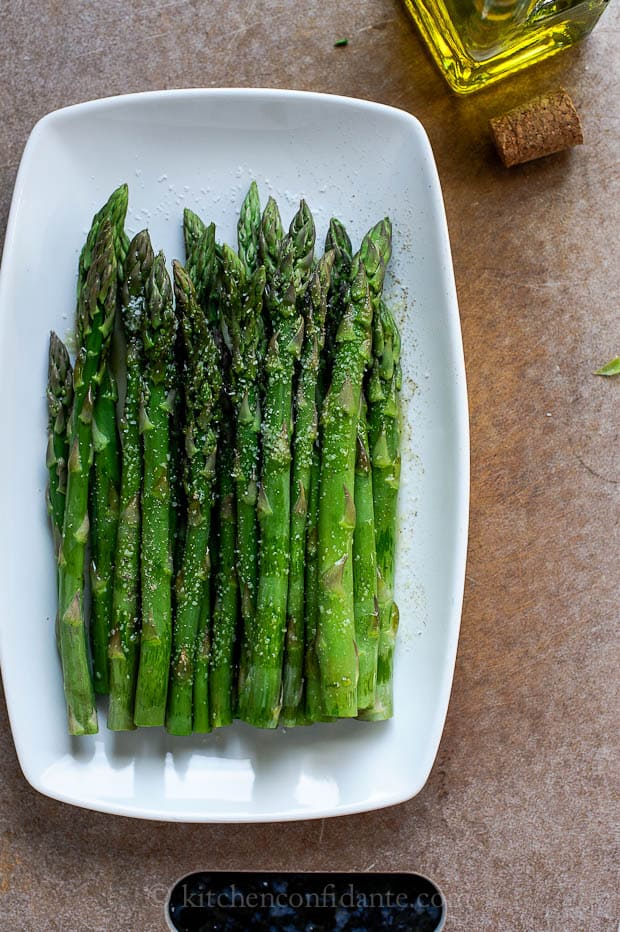 A white rectangular plate filled with raw asparagus that has been tossed with oil, salt, pepper, and vinegar. A bottle of oil and a cork sit beside the plate.