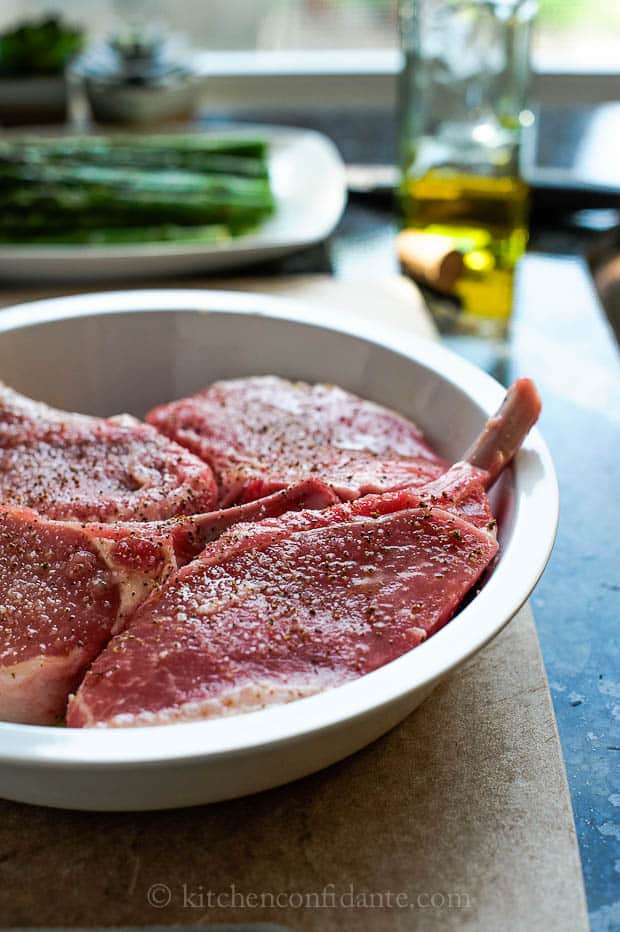 A bowl with three pork chops sprinkled with oil, vinegar, salt, and pepper sits on a wooden cutting board.