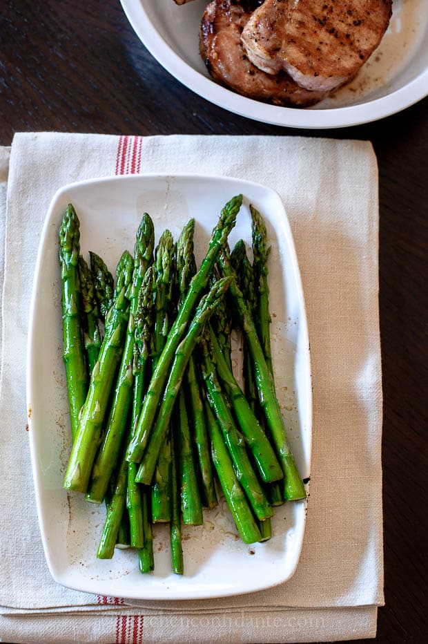 A plate of grilled asparagus sits on a white towel with a red stripe. A bowl of pork chops sits in the background.