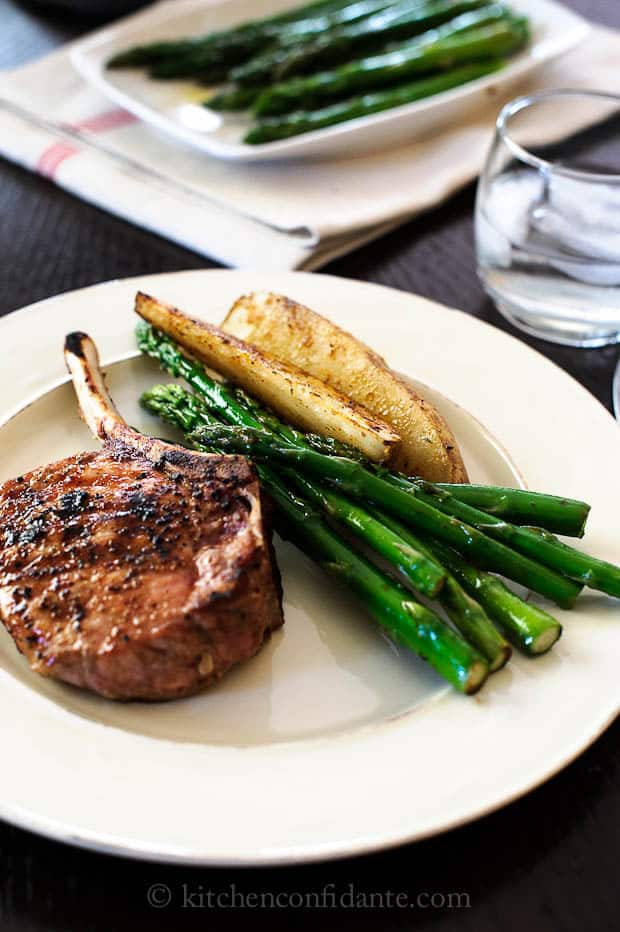 A dinner plate at a table with a pork chop, asparagus, and potato wedges. The serving plate sits in the background.