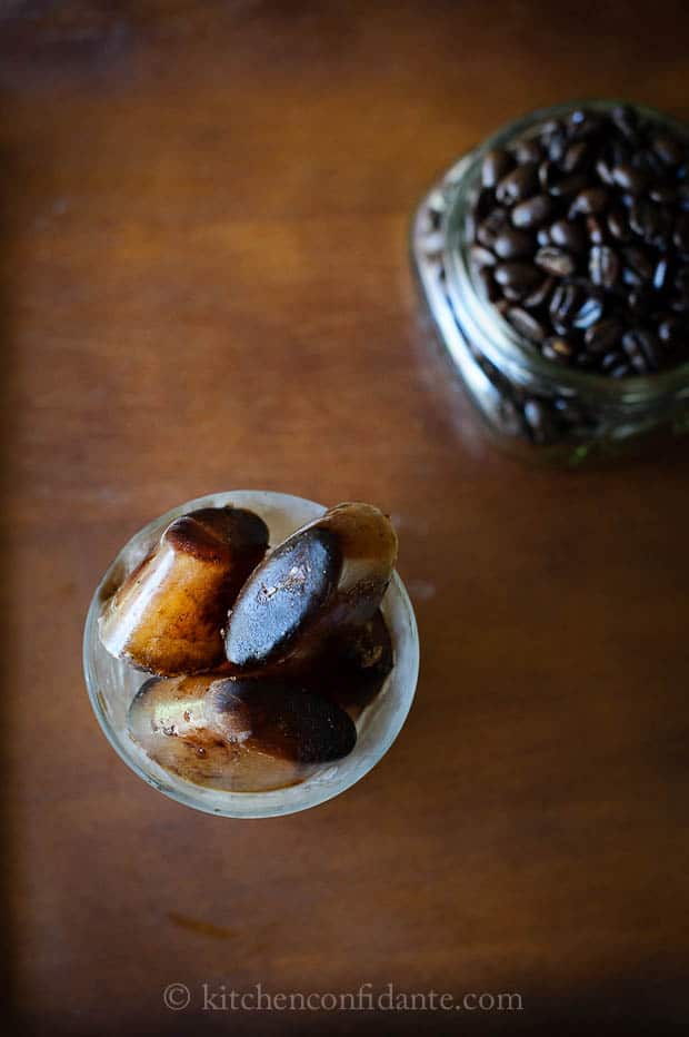 A glass filled with coffee ice cubes sits on a table with a jar of coffee beans in the background.