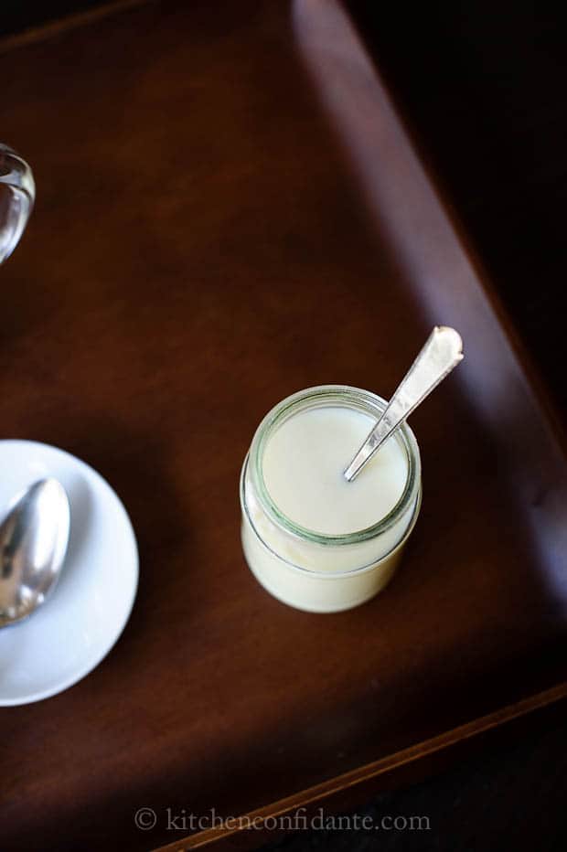 A jar filled with condensed milk with a spoon handle resting on the lip. A small plate and another spoon sit nearby.