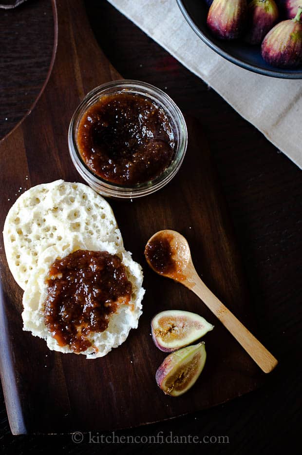 The balsamic fig jam recipe is completed, and a small wooden spoon sits on the table after applying a large smear of jam onto an english muffin.