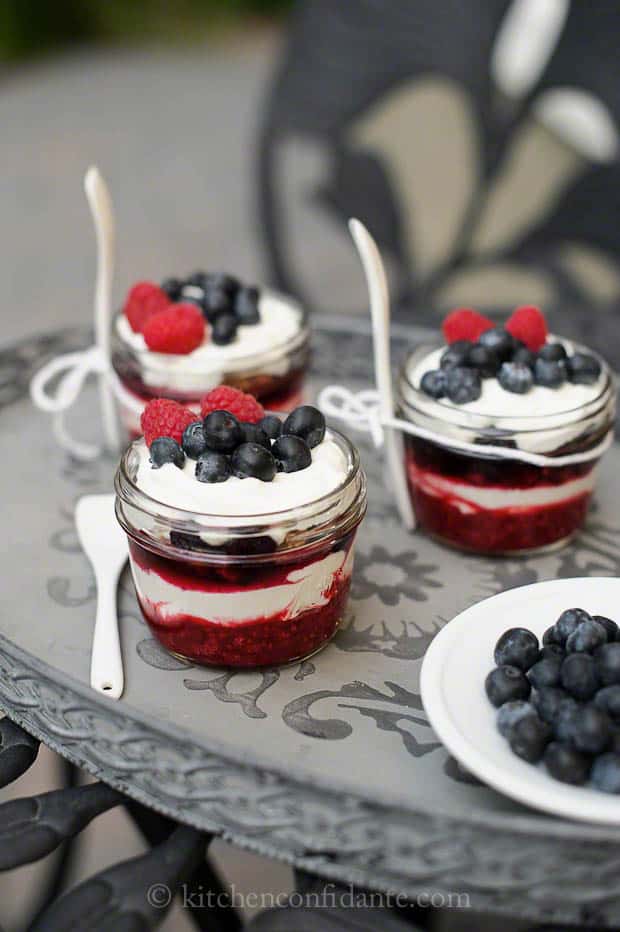 Three small jam jars sit on a tray. The jars are filled with layers of fruit, creamy filling, and a sweet crunch.