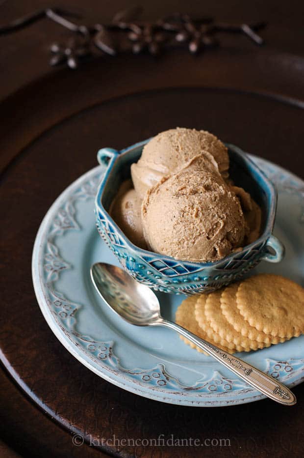 A blue dish filled with a light tea colored icecream sits on a saucer with a spoon and five coconut cookies.