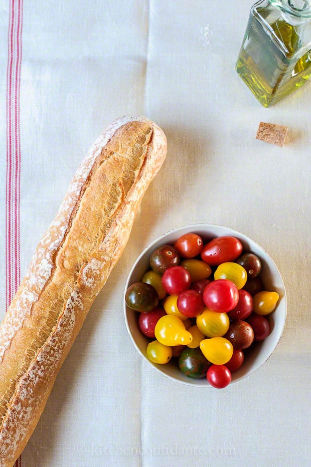 A baguette lies beside a bowl of yellow and red heirloom tomatoes. A bottle of olive oil and a cork lay in the background.