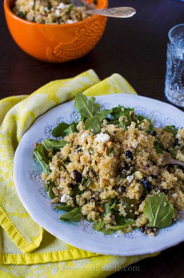 A blue bowl of quinoa black bean salad lies on a table. A fork is on the plate, and a cup of water and an orange bowl is in the background.