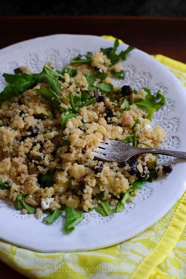 A plate of quinoa black bean salad sits on a yellow cloth. A fork lies on the plate, and corn, shallots, and arugala are mixed into the salad.