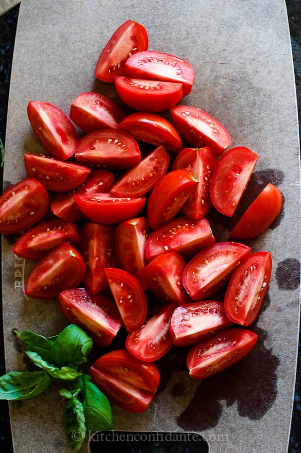 A gray cutting board full of freshly quarted roma tomatoes and fresh basil leaves ready to make homemade spaghetti sauce.