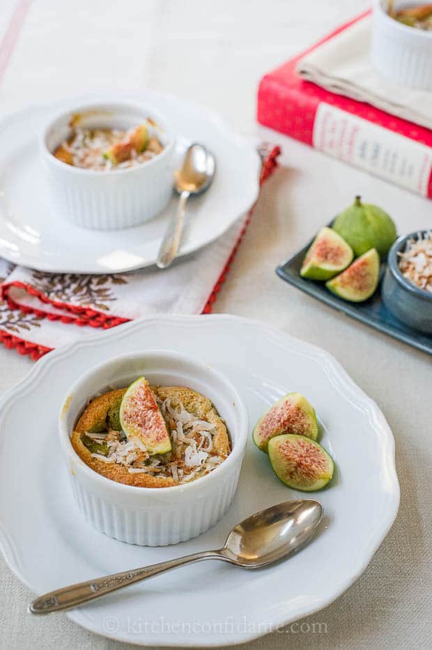 Two dishes of coconut fig clafoutis sit on individual plates, with spoons at the ready, and fresh figs for extra garnish.