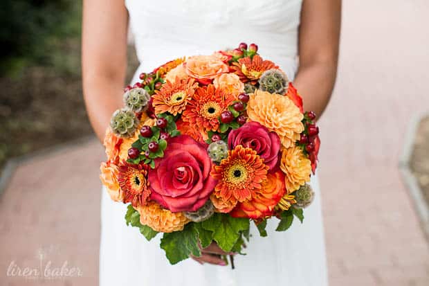 A bride in a white wedding dress holds a bright bouquet filled with red and orange summer flowers.