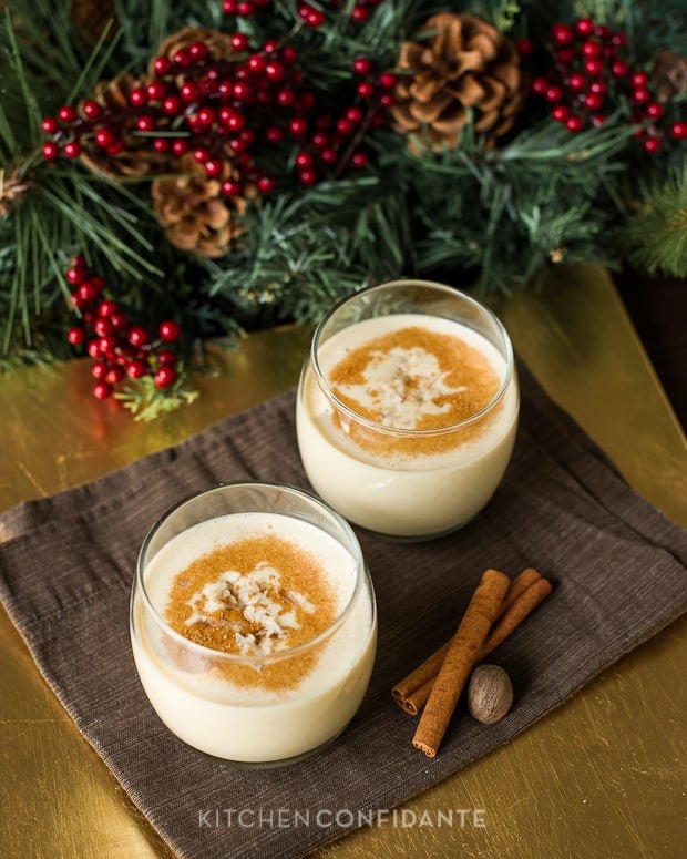 Two glasses of eggnog sitting on a brown napkin beside a hazelnut and cinnamon sticks.