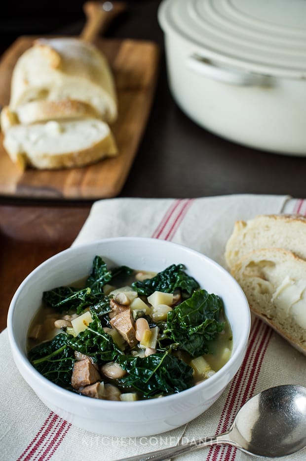 A white bowl of Kale Parsnip & Sausage Soup. Bread with butter sits beside the bowl as well as in the background.