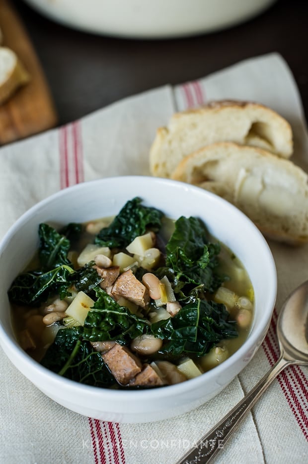 A white bowl of Kale Parsnip & Sausage Soup. The bowl sits on a white towel with red stripes. A spoon and two pieces of bread lay beside the bowl.