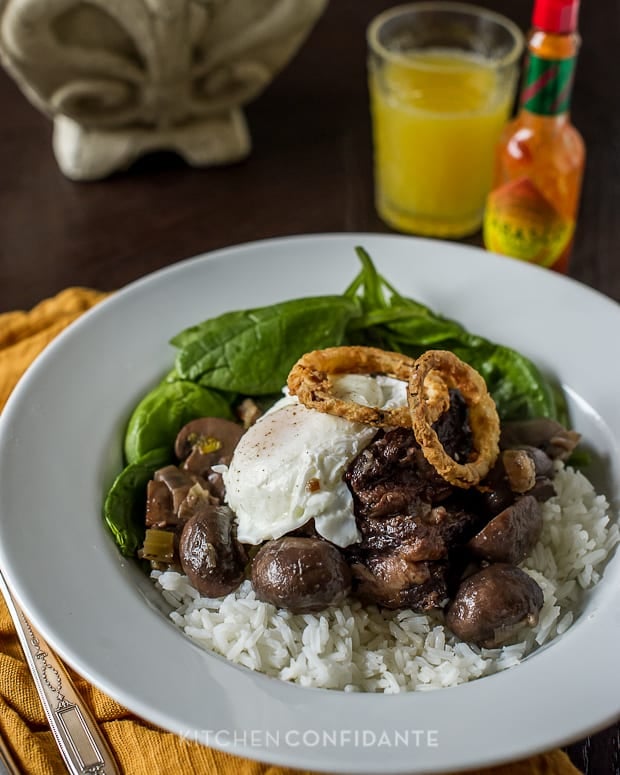 A white dish filled with Short Rib Loco Moco, with a bottle of tobasco sauce and a glass of orange juice in the background.