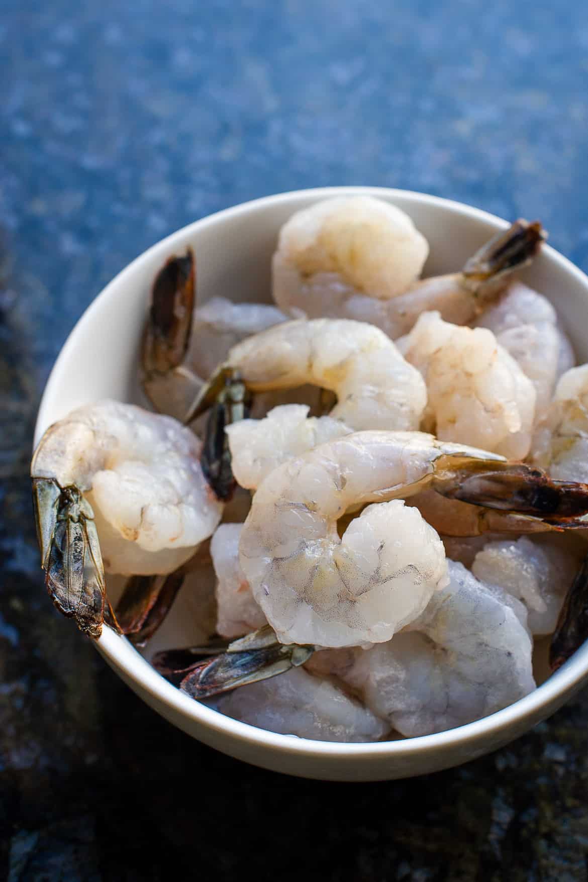 A white bowl of raw shrimp on a blue table.