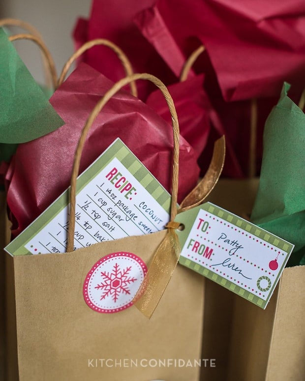 A brown paper bag with decorative red and white snowflake sticker. The bag is stuffed with red and green tissue paper, with a recipe card tucked inside, and a To/From tag attached to the handle.