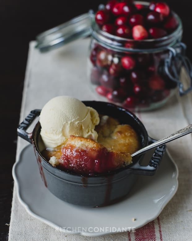 An individual Cranberry Rhubarb Cobbler baked in a small baking dish and a glass jar of cranberries.