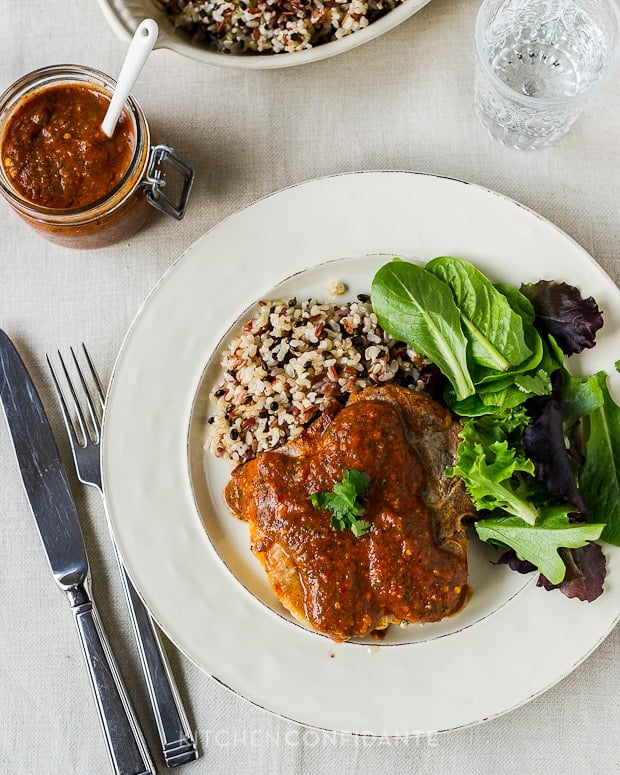 Fire Roasted Tomato Harissa on a grilled pork chop served on a white plate with mixed grains and a green salad.