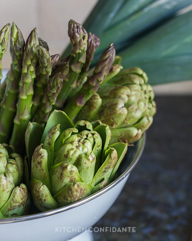 Fresh asparagus and artichokes in a white colander. 