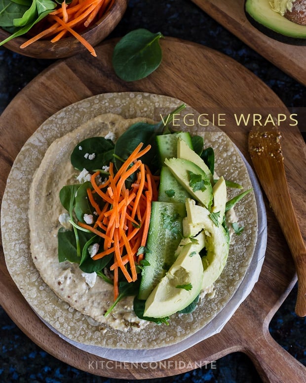 A whole grain tortilla topped with hummus, spinach, carrots, cucumber slices, and avocado slices with the words, "Veggie Wraps".