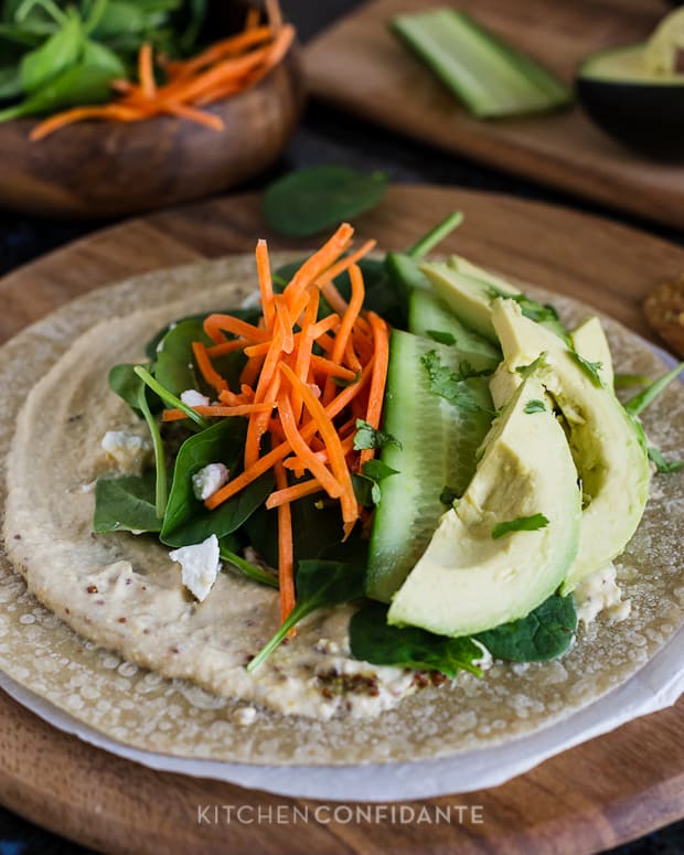 A homemade veggie wrap with a whole grain tortilla topped with hummus, spinach, carrots, cucumber slices, and avocado slices with the words.