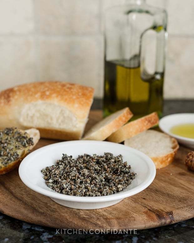 A bowl of Egyptian Macadamia Nut Dukkah, a loaf of sliced bread, and a bottle of olive oil.