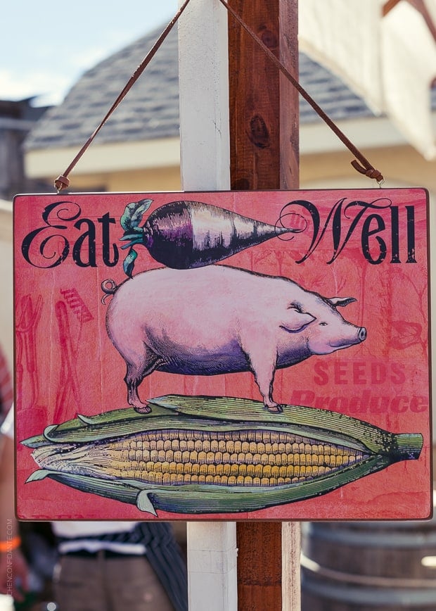 A pink sign that says "Eat Well". An image of a radish is on top of an image of a pig which is on an image of a partially husked ear of corn.