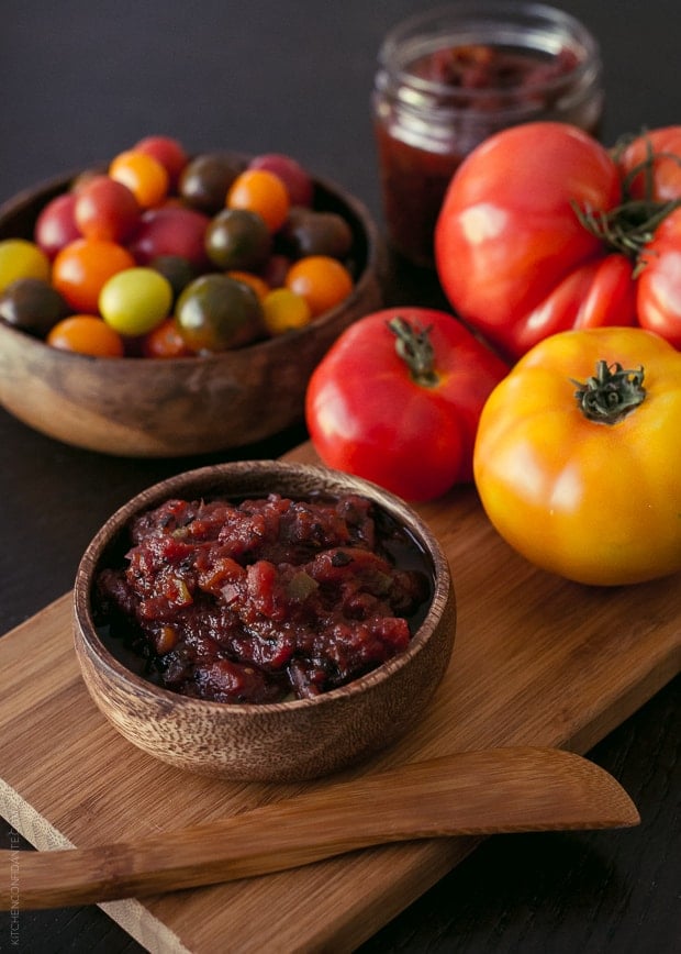 A wooden bowl filled with Fire Roasted Tomato Jam. In the backgroudn are tomatoes of various sizes and varied colors.