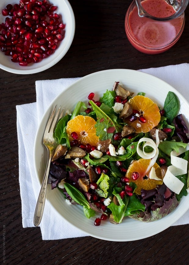 A plate of Chestnut Salad with Pomegranate Dressing, a bowl of pomegranate arils, and a small pitcher of pomegranate dressing.