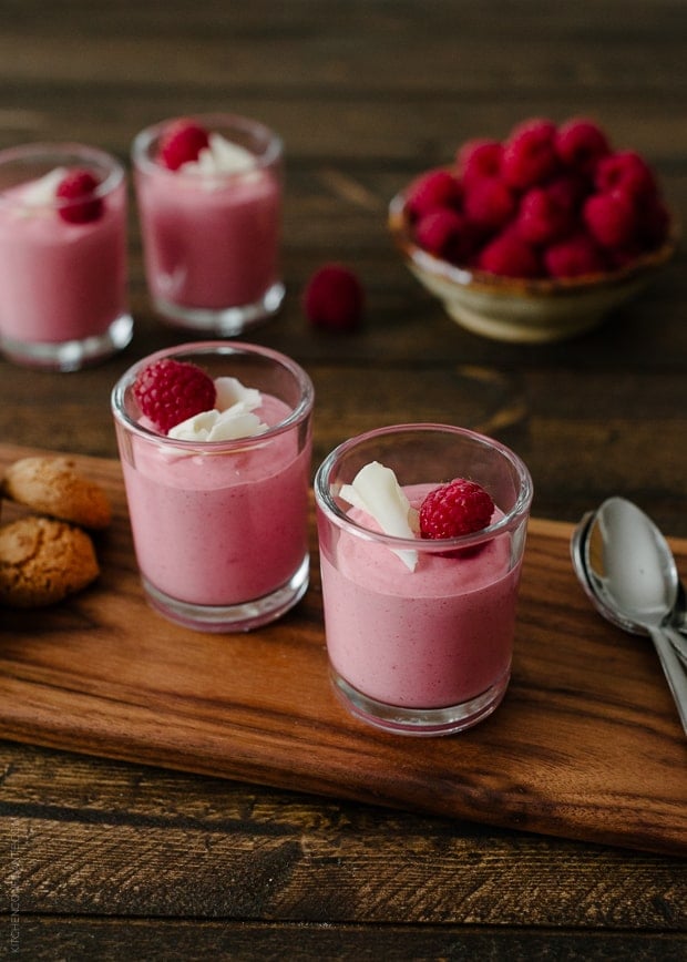 Small glass cups of raspberry mousse garnished with white chocolate and raspberries.