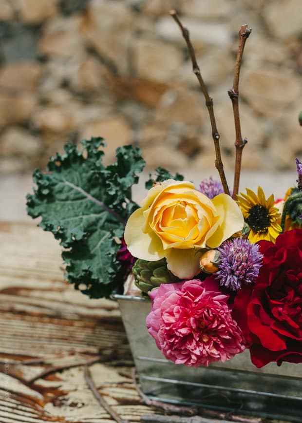 A metal container holding an arrangement of fresh flowers and vegetables.