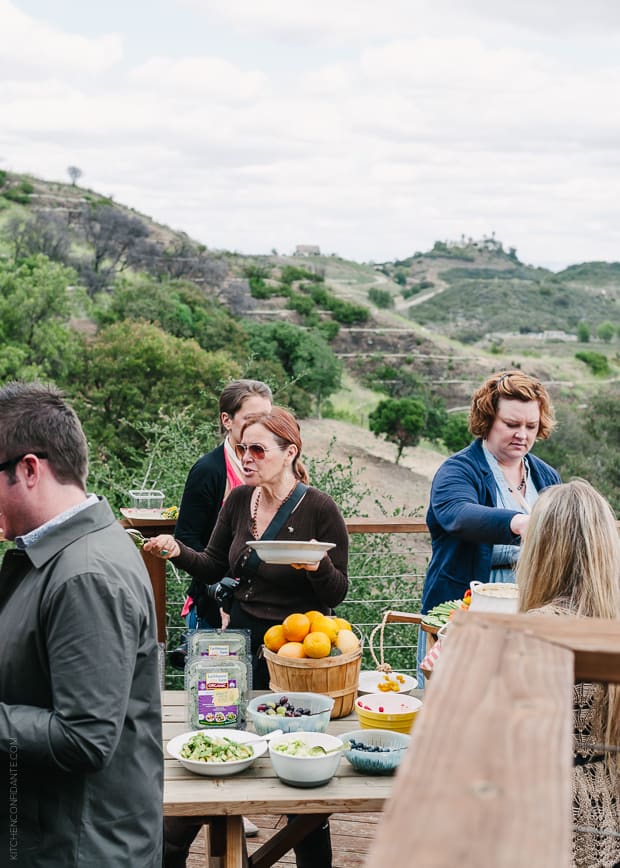 Various people filling plates at an outdoor serving table. Beautiful scenery and hills lay in the background.