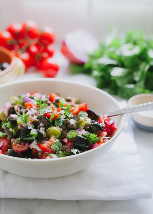 A white bowl piled with roughly chopped Tomato Olive Salsa. The ingredients to this recipe are in the background.