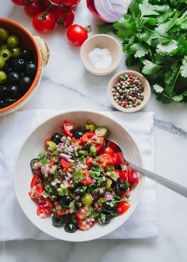 A bowl of Tomato Olive Salsa, with the fresh ingredients sitting in the background.