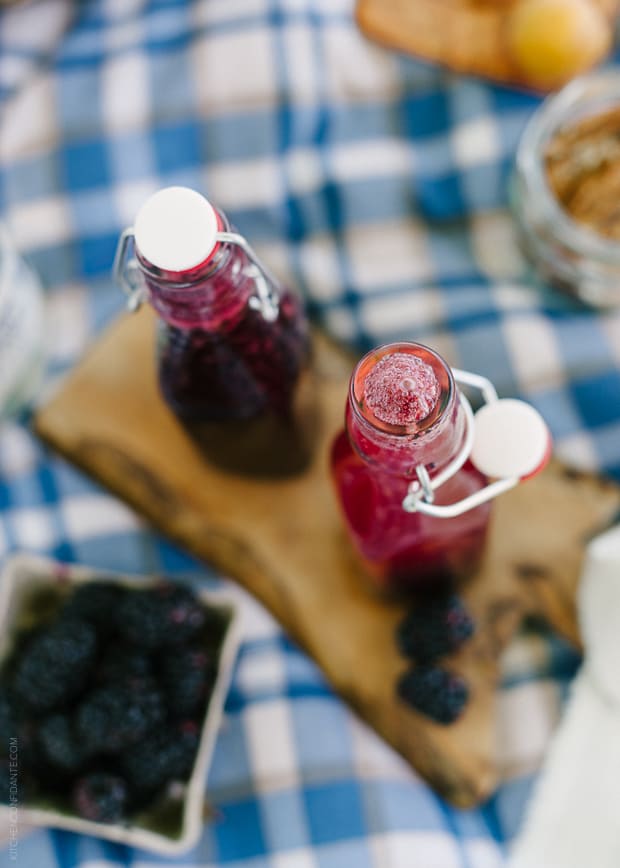 Blackberry Shrub Syrup in glass bottles surrounded by blackberries.